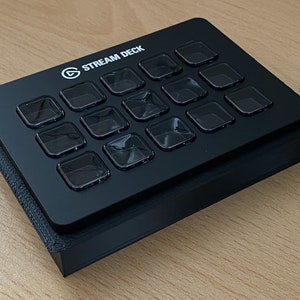 Könnte beinhalten: Ein schwarzes Stream Deck mit 15 Tasten, jede mit einer durchsichtigen Plastikabdeckung. Das Stream Deck ist ein Gerät, das für Live-Streaming und Content Creation verwendet wird.