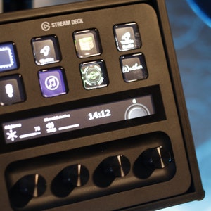 Puede incluir: Un Stream Deck negro con una pantalla digital que muestra la hora 14:12 y una perilla de volumen. El deck tiene varios botones con iconos para diferentes funciones, incluyendo un micrófono, una nota musical y una cámara.