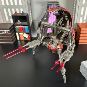 May include: A red and silver articulated robot toy with red laser beams. The robot is posed with its legs extended and its arms raised. The robot is sitting on a black surface.