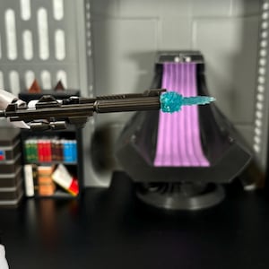 May include: A white action figure of a stormtrooper holding a black blaster rifle with a blue energy blast effect. The figure is posed in a firing stance. The background is a dark grey room with a purple and black throne.