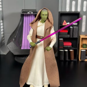 May include: An action figure of a green-skinned character from a science fiction franchise. The figure is dressed in a white gown and a brown hooded cloak, holding a purple lightsaber. The background is grey, with a black shelf unit.