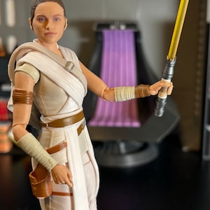 May include: A white action figure of a female character with brown hair and a white and brown outfit. The figure is holding a yellow lightsaber in her right hand. The figure is posed in a fighting stance.