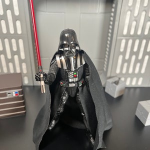 May include: Darth Vader action figure with a black cape and a red lightsaber. The figure is posed with the lightsaber raised in a fighting stance.