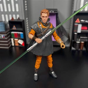 May include: A green lightsaber with a silver handle is held by a figure wearing a brown and black outfit. The figure has short brown hair and is wearing a brown belt with a silver buckle.