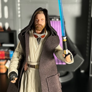 May include: A brown hooded robe with a white undergarment and a lightsaber. The figure is wearing goggles and has a beard.