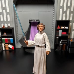May include: A white-robed action figure of Princess Leia from Star Wars, holding a blue lightsaber. The figure is posed in a standing position with her right hand holding the lightsaber and her left hand at her side. The figure is standing on a black surface in front of a grey and purple background.