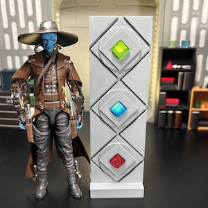 Op de afbeelding: Een witte muur met drie ruitvormige panelen, elk met een edelsteen in een andere kleur. De edelstenen zijn groen, blauw en rood. Een blauwhuidige action figure met een bruin leren jack en een brede hoed staat voor de muur.