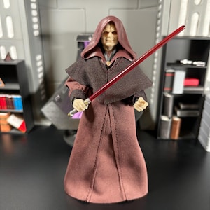 May include: A detailed action figure of a character from a science fiction franchise. The figure is dressed in a maroon robe and hood, holding a red lightsaber. The figure has a stern expression and is posed in a room with futuristic elements.