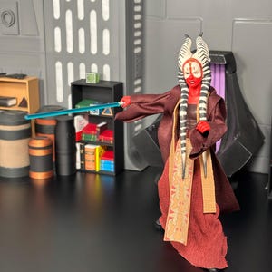 May include: A red-skinned action figure of a Twi'lek Jedi with a blue lightsaber, wearing a maroon robe and a patterned sash. The figure has white and black head tails and is posed in a sci-fi setting with props.