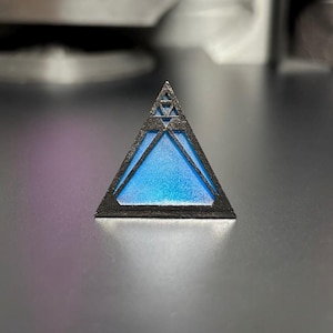 May include: A small, black triangular prism with a blue, iridescent centre. The prism has a geometric design on the front.
