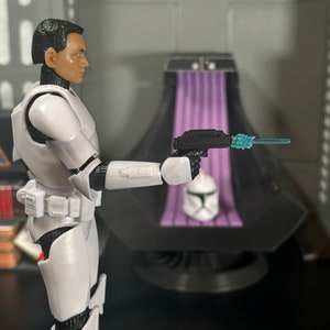 May include: A white action figure of a stormtrooper holding a black blaster with a blue tip. The figure is standing in front of a purple and black throne.