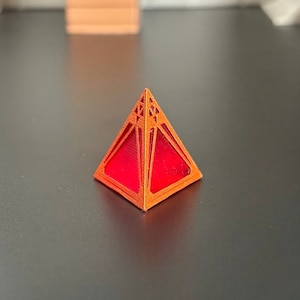 Puede incluir: Una pirámide geométrica roja y naranja con intrincados recortes. La pirámide está hecha de un material translúcido, que permite que la luz brille a través de ella.
