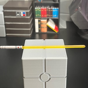 May include: A silver and yellow lightsaber rests on a gray, geometric stand. The lightsaber has a detailed handle with red accents and a bright yellow blade. The background includes a shelf with books and other decorative items.