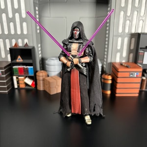 May include: A black and red action figure of a Star Wars character with a purple lightsaber. The figure is wearing a black hooded robe and a mask.