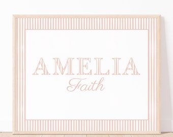 May include: A framed print with the name "AMELIA" in a light pink, decorative font above the word "Faith" in a cursive script. The print has a striped border in light pink and white, set against a white background.