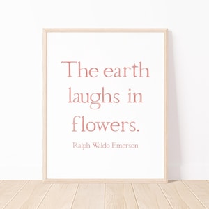 May include: A framed print featuring the quote "The earth laughs in flowers" in a soft pink font. The quote is attributed to Ralph Waldo Emerson. The frame is light wood, and the background is white.