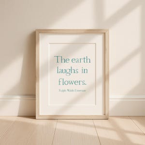 May include: A framed print featuring the quote "The earth laughs in flowers" in teal lettering, attributed to Ralph Waldo Emerson. The frame is light wood, and the print has a white mount.
