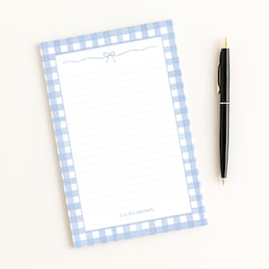 May include: A lined notepad with a light blue gingham border and a bow illustration at the top. The notepad has the name "LAURA BROWN" printed at the bottom. A black pen with gold accents rests beside the notepad.