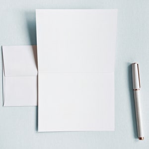 Peut inclure: Une carte blanche avec un bord pli&eacute; et une enveloppe blanche. Un stylo blanc avec un capuchon dor&eacute; est pos&eacute; sur une surface bleu clair.