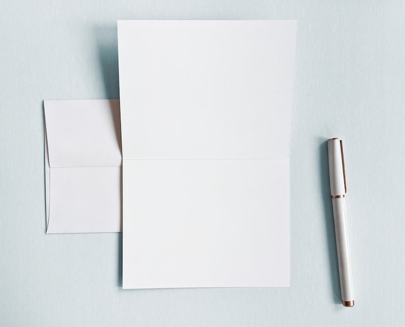 Peut inclure: Une carte blanche avec un bord pli&eacute; et une enveloppe blanche. Un stylo blanc avec un capuchon dor&eacute; est pos&eacute; sur une surface bleu clair.