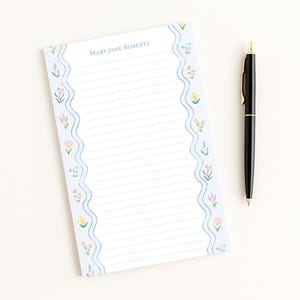 May include: A lined notepad with a light blue floral border and the text "MARY JANE ROBERTS" at the top. A black pen with gold accents rests beside the notepad on a white surface. The notepad features a wavy border design.