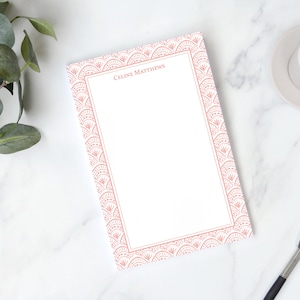 May include: A white notepad with a decorative coral border and the name "Celine Matthews" printed at the top. A cup of coffee and a pen are next to the notepad on a marble surface. Green leaves are in the upper left corner.