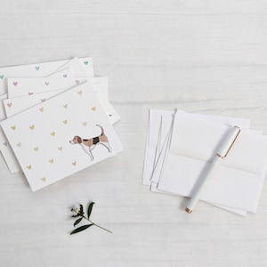 Peut inclure: Une pile de cartes de v&oelig;ux vierges avec une illustration de chien et une dispersion de c&oelig;urs color&eacute;s. Les cartes sont sur une surface en bois blanc avec un stylo blanc et une petite branche de verdure.