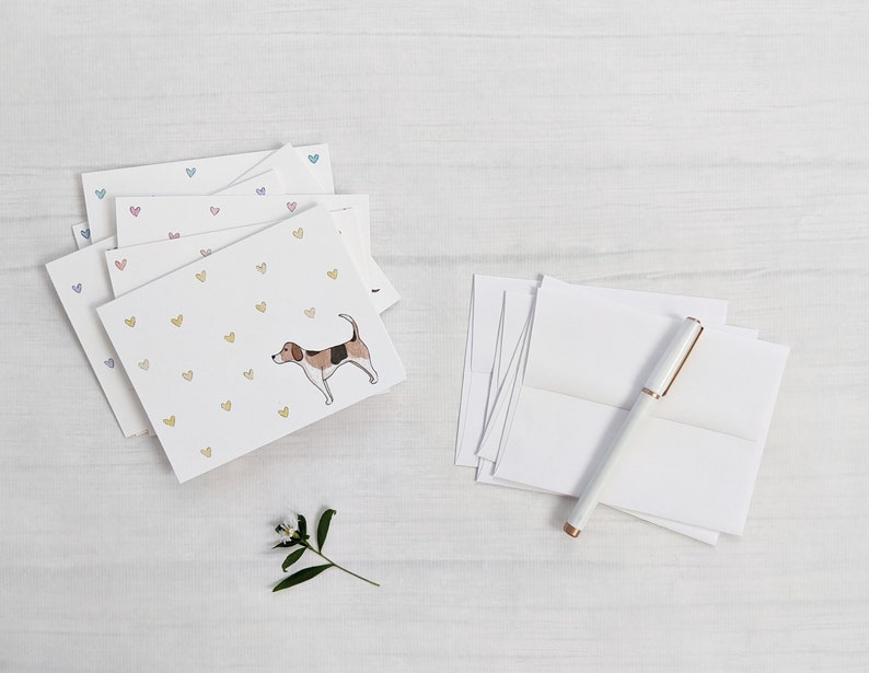 Peut inclure: Une pile de cartes de v&oelig;ux vierges avec une illustration de chien et une dispersion de c&oelig;urs color&eacute;s. Les cartes sont sur une surface en bois blanc avec un stylo blanc et une petite branche de verdure.