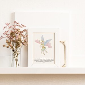 May include: A framed watercolor print of an angel with blue and pink wings, holding a staff. The print is in a white frame, next to a small gold corner piece and a vase of dried pink flowers. The background is white.