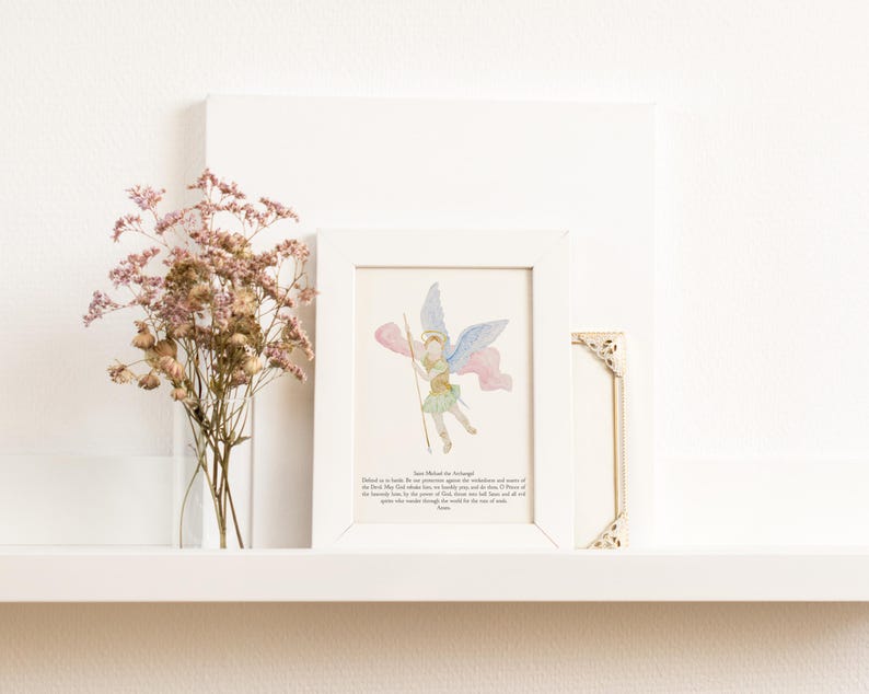May include: A framed watercolor print of an angel with blue and pink wings, holding a staff. The print is in a white frame, next to a small gold corner piece and a vase of dried pink flowers. The background is white.