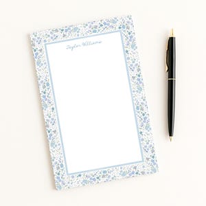 May include: A white notepad with a blue floral border and the name "Taylor Williams" printed in cursive. A black pen with gold accents rests beside the notepad. Stationery.