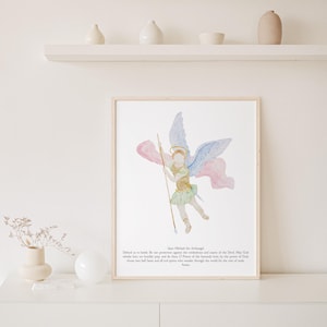 May include: Watercolor print of Saint Michael the Archangel, framed in a light wood. The artwork depicts the angel with blue and pink wings, holding a spear. Text at the bottom includes a prayer. The artwork is displayed on a white surface.