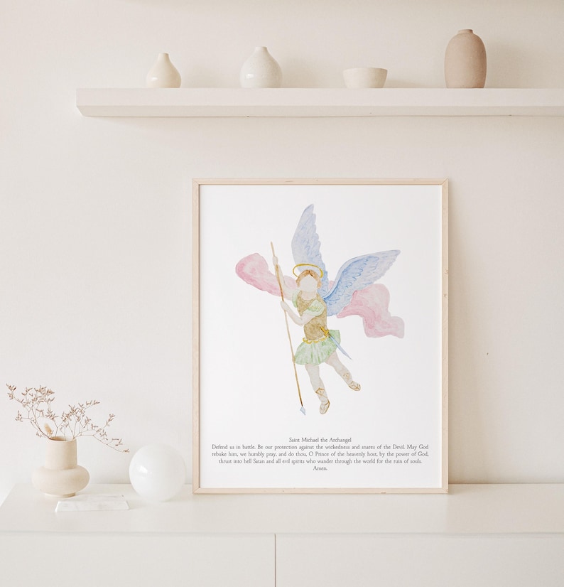 May include: Watercolor print of Saint Michael the Archangel, framed in a light wood. The artwork depicts the angel with blue and pink wings, holding a spear. Text at the bottom includes a prayer. The artwork is displayed on a white surface.
