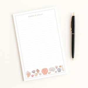 May include: A rectangular notepad with horizontal lines and a border featuring watercolor seashells at the bottom. The name Madeline R. Johnson is printed at the top. A black ballpoint pen with gold accents is positioned to the right of the notepad.