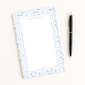 May include: A notepad with a floral border and the text "Vivienne Walton" is next to a black pen with gold accents. The notepad has a light blue border and floral design. The pen is approximately 5.5 inches long.
