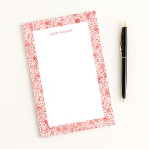 May include: A floral notepad with a red and pink rose pattern border and the name "Jamie Wilson" printed at the top. A black pen with gold accents rests beside the notepad on a white surface.