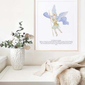 May include: A framed watercolor print of Saint Michael the Archangel, depicted with wings, a spear, and a halo. The artwork features soft blues, greens, and gold tones. Text at the bottom reads, "Defend us in battle..." A decorative vase and a folded cream-colored blanket are also visible.
