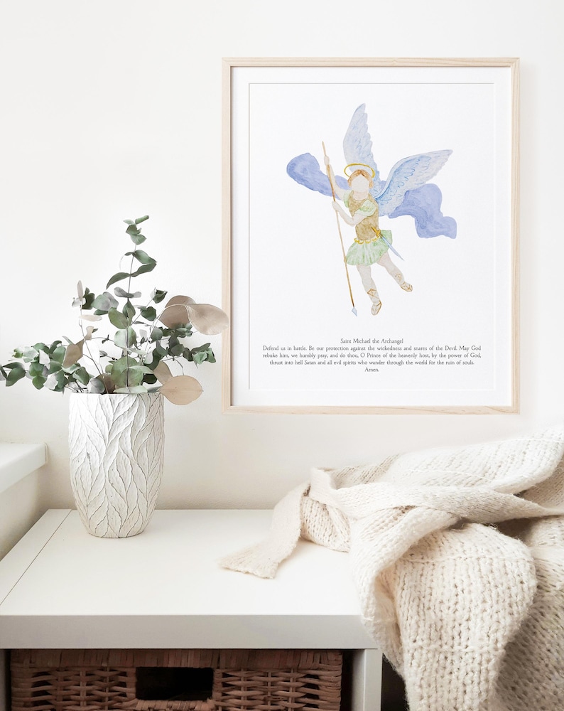 May include: A framed watercolor print of Saint Michael the Archangel, depicted with wings, a spear, and a halo. The artwork features soft blues, greens, and gold tones. Text at the bottom reads, "Defend us in battle..." A decorative vase and a folded cream-colored blanket are also visible.