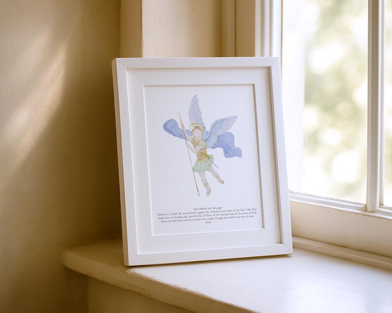 May include: A framed watercolor print of Archangel Michael, depicted with blue wings and a spear. The artwork is in a white frame and includes text at the bottom. The print is displayed on a white windowsill.