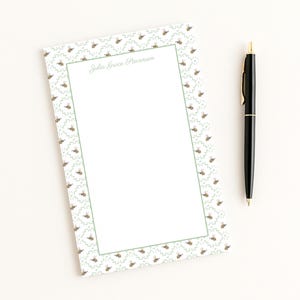 May include: A white notepad with a green border and a repeating pattern of bees and leaves. The notepad has the name "Julia Grace Stevenson" printed at the top. A black pen with gold accents rests beside the notepad.