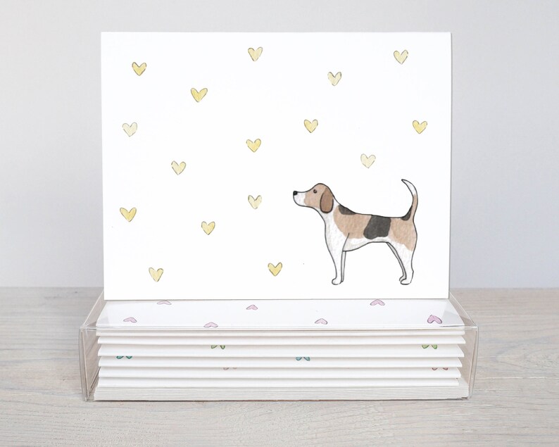 Peut inclure: Un ensemble de 8 cartes de v&oelig;ux vierges illustrant un beagle en aquarelle sur fond de c&oelig;urs jaunes. Les cartes sont emball&eacute;es dans une bo&icirc;te en plastique transparente avec un motif de c&oelig;urs roses et verts.