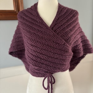 May include: A purple crocheted wrap with a V-neck and a tie at the bottom.