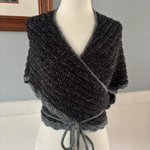 May include: A black and gray crocheted wrap with a tie closure. The wrap is made with a textured yarn and has a scalloped edge.