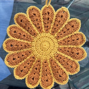 May include: A handmade crochet sunflower trivet in shades of yellow and brown. The trivet features a circular center with radiating petals, each outlined in a darker brown. A small loop is attached for hanging. The trivet is designed to protect surfaces from heat.