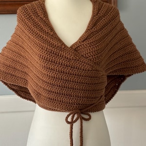 May include: A brown crocheted shawl with a V-neck and a tie at the bottom.