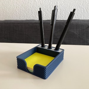 Könnte beinhalten: Ein marineblauer Schreibtisch-Organizer mit drei schwarzen Stiften und einem Stapel gelber Haftnotizen. Der Organizer hat ein strukturiertes, geriffeltes Design und eine rechteckige Form mit abgerundeten Ecken. Die Stifte stehen aufrecht im Organizer.
