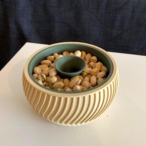 Könnte beinhalten: Eine beige-grüne Servierschale gefüllt mit Pistazien. Die Schale hat einen zentralen Dip-Bereich und ein geriffeltes Außendesign. Die Pistazien sind in ihren Schalen, wobei einige die grüne Nuss im Inneren zeigen.