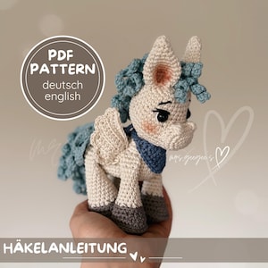 May include: A crocheted unicorn with cream-colored body, gray hooves, and blue mane and tail. The unicorn has wings and a blue scarf. The image includes the text "PDF PATTERN deutsch english" and "HÄKELANLEITUNG".