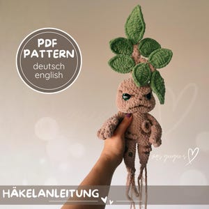 Könnte beinhalten: Gehäkeltes Mandrake-Pflanzenspielzeug mit hellbraunem Körper, grünen Blättern und einem strengen Gesichtsausdruck. Das Spielzeug hat eine detaillierte Wurzelstruktur und wird von einer Hand gehalten. Der Text "PDF PATTERN deutsch english" und "HÄKELANLEITUNG" ist sichtbar.