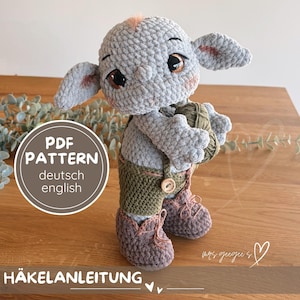 May include: A handmade crochet doll in a whimsical style. The doll is gray with large brown eyes, pink accents, and wears green overalls and brown shoes. It holds a ball of yarn. The image includes text that says "PDF PATTERN deutsch english" and "HÄKELANLEITUNG".